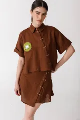 Camisa corta marrón de manga corta, con doble cartera diagonal con botones de madera, cuello clásico y botamangas marcados. Aplique de crochet hecho a mano con forma de kiwi en el bolsillo.