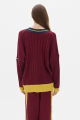 Sweater tejido de punto color crema, con cuello en V con borde negro y doble línea blanca, mangas largas con puños negros, diseño con trenzas en el frente y corte oversize.