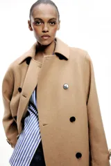 Abrigo color camel confeccionado en hilatura con lana. Tiene cuello solapa, manga larga, bolsillos delanteros ocultos en costura y cierre frontal cruzado con botones negros.