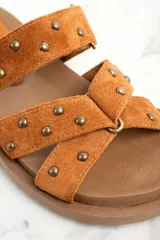 Sandalias marrones de gamuza con apliques de tachas doradas, con dos tiras anchas sobre el empeine y suela plana.