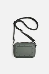 Bandolera rígida color verde militar, con formato de caja rectangular y detalles de remaches. Tiene un compartimento principal con cierre de cremallera y un bolsillo interior plano. Incluye asa de hombro ajustable y extraíble.