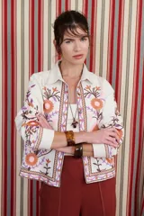 Chaqueta de tejido color crema con cuello camisero y bordados florales en tonos naranja, rosa y morado. Presenta cierre frontal con botones y puños anchos.