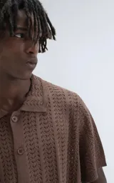 Camisa de tejido de punto color café, manga corta y cuello polo con botones frontales. Presenta un patrón calado de espiga o chevron.