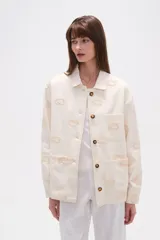Campera de algodón color crudo, estilo camisa o chaqueta ligera, con bordados en forma de paisley en tono beige claro. Posee cuello camisero, cierre frontal con botones marrones y bolsillos delanteros.