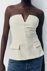 Top blanco de escote recto con detalle de pieza metálica en forma de V forrada y hombros descubiertos. Tiene falsos bolsillos con solapa en el delantero, forro interior combinado y cierre lateral con cremallera oculta en la costura.