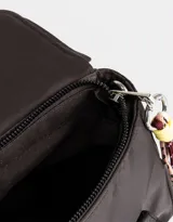 Cartera tipo city color beige, de tela deportiva, con doble asa corta, correa regulable desmontable, solapa con broche a presión y charm colgante.