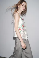 Top blanco de cuello mao y sin mangas, con bordados florales en el frente y cierre con botones.