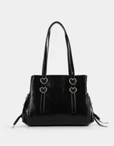 Cartera tipo city de cuerina color negro con efecto charol. Tiene manijas superiores con apliques metálicos en forma de corazón, cierre superior y bolsillos laterales con trenzado decorativo.