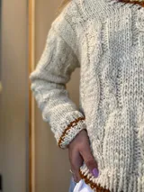 Sweater artesanal de lana color crudo con diseño de trenzas y ochos. Tiene cuello redondo y terminaciones en marrón.
