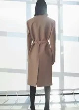 Abrigo largo color beige con cuello de solapa, hombros marcados, cinturón con hebilla metálica, bolsillos laterales con solapa y abertura en la parte posterior del bajo.