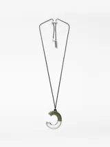 Collar con cordón largo y regulable de poliéster negro. El dije es una pieza irregular con forma de 'C' o media luna, mitad de resina verde y mitad de metal plateado. El ajuste del cordón es mediante un deslizador metálico con textura de sierra.