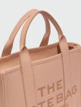 Bolso tote de cuero negro Marc Jacobs, tamaño mediano, con doble asa corta y correa larga extraíble. Estampado frontal con el texto "THE TOTE BAG" y el nombre de la marca.