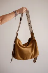 Bolso de hombro tipo hobo de gamuza color marrón óxido, con correa ancha ajustable de color negro y herrajes metálicos plateados. El nombre de la marca "GANESHA" está grabado sutilmente en el cuerpo del bolso.
