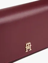 Cartera rectangular de color borgoña con acabado liso y solapa frontal con cierre magnético. Presenta un monograma TH en relieve en la parte delantera. Incluye correa ajustable y extraíble para llevar al hombro o como cartera de mano.