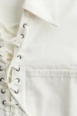 Vestido corto de denim color blanco, con cuello camisero, mangas cortas, dos bolsillos frontales y ajuste con cordones cruzados en el escote.