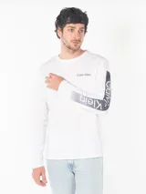 Remera blanca de algodón con cuello redondo y manga larga. Logo de Calvin Klein estampado en el pecho y diseño con letras en la manga.