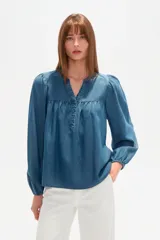 Blusa de lyocell en tono azul denim, con cuello en V, cierre frontal parcial con botones y mangas largas abullonadas con puños elásticos. Presenta un diseño holgado con detalle de fruncido en el pecho.