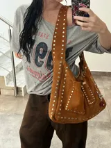 Cartera bandolera marrón de cuero con remaches metálicos plateados. Tiene correa ancha regulable, bolsillo externo y argolla para colgar accesorios.