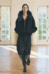 Chaqueta negra de cuello redondo confeccionada en hilatura con 50% lana. Tiene capucha extraíble, manga larga con puño interior, bolsillos delanteros y bajo ajustable con cordón elástico. Cierre frontal con cremallera metálica.