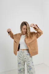 Chaqueta color beige de corte recto, estilo camisa, confeccionada en gamuza con elastano. Presenta cuello camisero, cierre frontal con botones metálicos, bolsillos grandes en el frente y charreteras en los hombros.