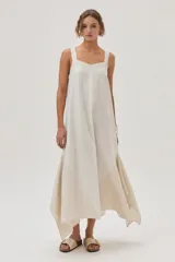 Vestido largo color crema, con breteles anchos y escote recto. Presenta corte irregular en el ruedo y volado en el bajo.