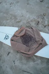 Hoodie marrón con capucha y cordón ajustable, con estampado en la espalda de un paisaje con palmeras, montañas y el texto "Glassy Waves".