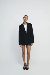 Blazer negra de corte clásico, con cuello de solapa y escote en pico. Tiene mangas largas con hombreras, bolsillos con solapa en el delantero y un bolsillo de vivo en el pecho. Está forrada y se cierra con un botón frontal.