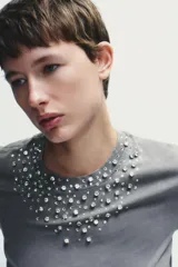 Camiseta gris de algodón con efecto lavado, cuello redondo y mangas cortas. Decoración con apliques de strass plateados en el escote.