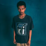 Remera negra de corte oversize con efecto lavado y estampado frontal con un diseño gráfico que incluye un logo con las letras 'KI', un dibujo de una casa y el número '420'.