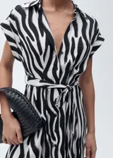 Vestido camisero midi con estampado de cebra en blanco y negro, cuello camisero, mangas cortas, cierre frontal con botones y lazo ajustable en la cintura.