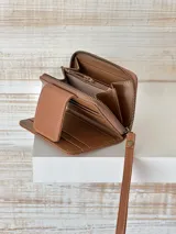 Billetera marrón de cuero con textura croco, cierre de cremallera y correa de muñeca. Presenta un broche con el logo de un búho.