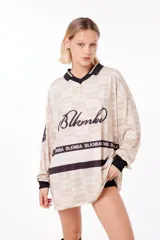 Camiseta oversize de poliéster color crema con diseño a cuadros, cuello en V con ribete negro, mangas largas con puños negros, logo "Blkmba" en el pecho y escudo "Mamba Soccer" en la manga.