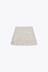 Falda pantalón corta color crudo con estampado de lunares negros, tiro medio, detalle de pliegues y volantes, y cierre lateral con cremallera.