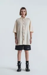 Camisa oversize color crema con cuello clásico, cierre frontal con botones marrones y mangas cortas.