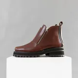 Bota de cuero vacuno color marrón, con interior forrado en corderito natural. Tiene suela de goma artesanal, punta cuadrada, cierre YKK en ambos lados y caña de 11cm.