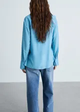 Camisa celeste de tejido fluido con cuello camisero, cierre frontal con botones y mangas largas con puños abotonados.