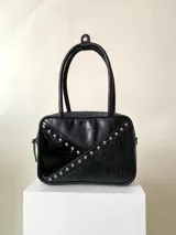 Bolso de mano rígido color negro, con textura símil cuero de cocodrilo y apliques de tachas plateadas. Tiene doble asa corta, cierre superior con cremallera y correa larga extraíble.