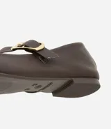 Zapatilla tipo bailarina de cuero color marrón, con punta redondeada y tira sobre el empeine con hebilla metálica dorada. Posee un tacón bajo de 1 cm y diseño clásico.