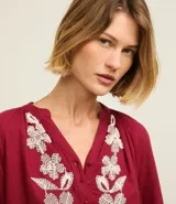 Blusa color borgoña de batista con cuello en V y botones. Presenta bordados florales blancos en el frente y las mangas largas con puños elásticos.