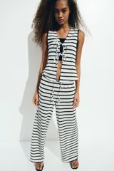 Top sin mangas de punto fino a rayas horizontales blancas y negras, con escote en V y cierre con lazo en el centro. Ribete festoneado en sisas y cuello.