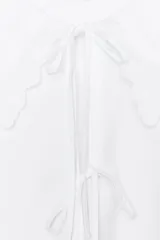 Camisa blanca de popelín con cuello bobo bordado y lazada. Tiene mangas abullonadas por debajo del codo con elástico y pinzas en la espalda. El cierre frontal es con lazadas.