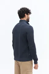 Buzo de algodón color verde musgo, con cuello alto y cierre frontal parcial. Presenta puños y cintura acanalados, y un pequeño logo bordado en el pecho.