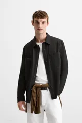 Sobrecamisa negra de tejido con elasticidad, cuello solapa y manga larga acabada en puño con botón a presión. Tiene bolsillos de plastrón en el pecho y cierre frontal con botones a presión.