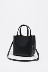Bolso pequeño tipo tote o cubo de piel vacuna texturizada color negro, con asas cortas superiores y correa larga ajustable para llevar al hombro o cruzado. Presenta el logo metálico de Bimba y Lola en dorado en el frente.