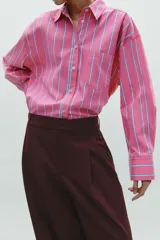 Camisa oversize rosada con rayas verticales blancas y celestes, cuello solapa, manga larga con puños abotonados, bolsillo de plastrón en el pecho y pinza en la espalda.