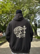 Canguro negro con capucha y cordones ajustables. Estampa en la espalda con el texto "Chema Surf Trip" y dibujos de un sol, una tabla de surf, una carpa y otros elementos relacionados con el surf.