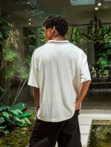 Chomba de tejido de punto cerrado color blanco, con cuello polo y mangas cortas. Presenta un bordado sutil en el frente y tres franjas negras en los hombros.