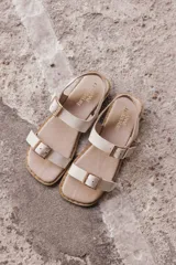 Sandalia plana de cuero color beige claro, con dos tiras anchas ajustables con hebillas metálicas cuadradas de color dorado. El borde de la plantilla está decorado con tachas metálicas doradas.