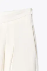 Pantalón blanco de tiro alto con pinzas delanteras, bolsillos laterales y pernera ancha. Cierre lateral con cremallera oculta.