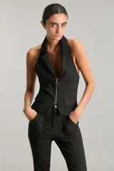 Chaleco negro con cuello halter y solapas sastreras. Cuenta con cierre frontal con cremallera metálica, espalda cavada y lazo de ajuste con avío metálico. Confeccionado en bengalina con efecto metalizado.